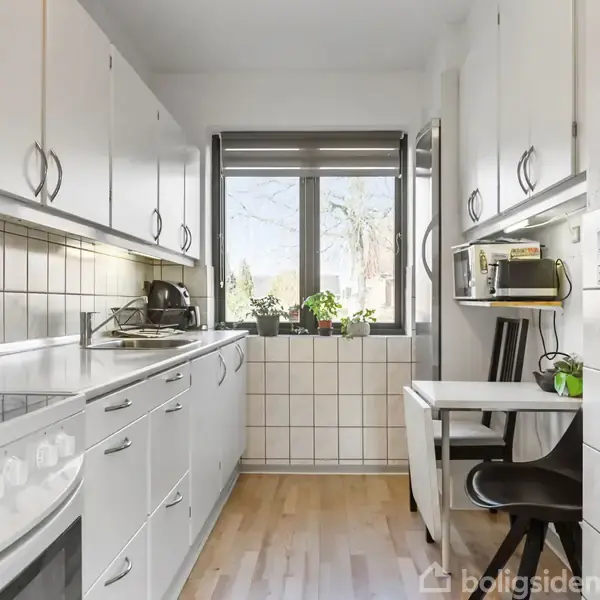 En lang, smal køkken med hvide skabe og trælamingulv. På bordpladen står en knivblok. Et bord ved vinduet har en mikrobølgeovn og plante. Ingen tekst er synlig.