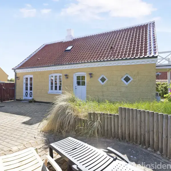 Et gult hus med rødt tegltag står i en solrig gårdsplads med fliser. Foran huset er der liggestole og en lav træhegn omgiver området.