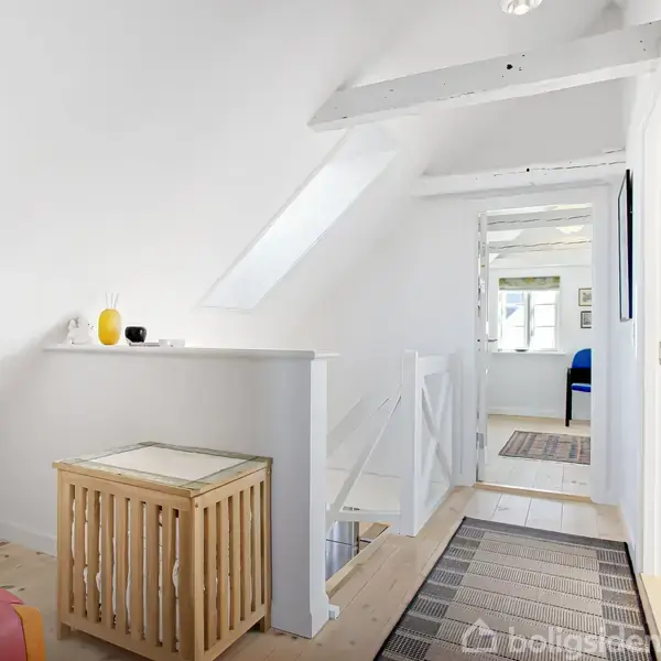 Et lyst, hvidmalet gangområde viser en trappe med en beskyttende halv væg. Et lille bord med kurv står ved siden af. Døren åbner til et rum med vindue.