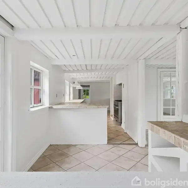 Et lyst, rummeligt køkken med hvide lofter og vægge har en bar og adskillige vinduer, der giver masser af lys. Der er store fliser på gulvet og en åben adgang til en terrasse.