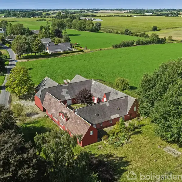 Rød gård i firkantet arrangement omgivet af grønne marker og træer. Beliggende ved en landevej med andre ejendomme synlige i baggrunden under en blå himmel.