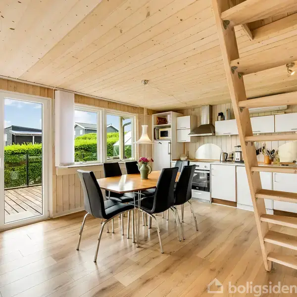 Et lyst køkken-alrum med træloft og -gulv. Et spisebord med sorte stole står centralt. Stige fører til hems. Store vinduer viser udsigt til terrasse og grøn have.