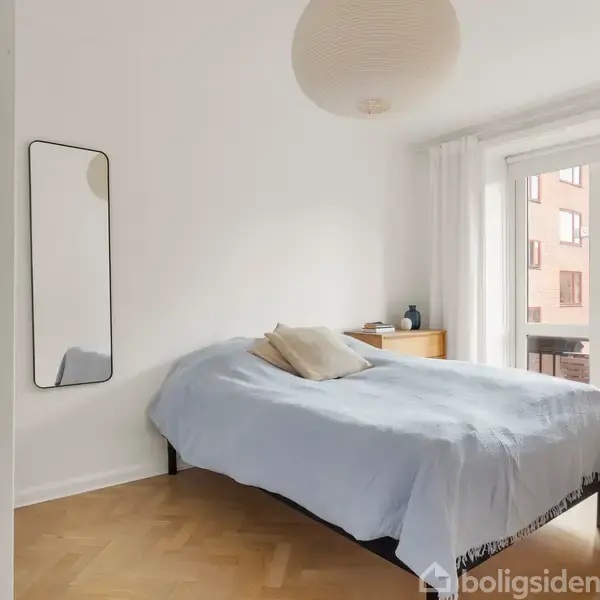 En seng dækket med et lyseblåt tæppe står i et lyst soveværelse. Ved siden af er et gulvspejl og en kommode. Store vinduer viser bygninger udenfor.