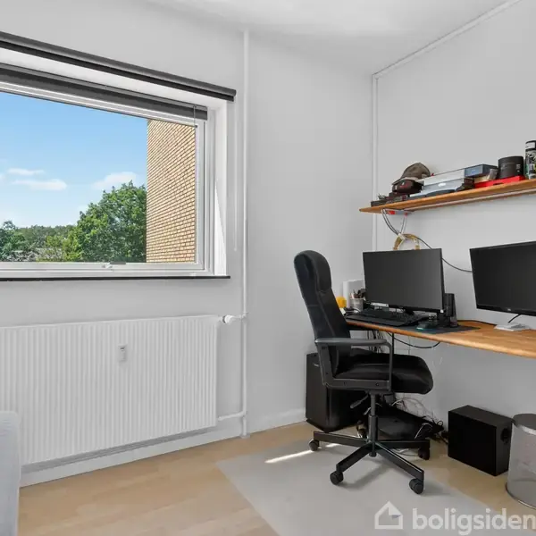 Et arbejdsbord står i et lyst rum med en kontorstol, to computerskærme og hylder med småting på væggen. Et vindue viser udsigt til en murstensbygning og træer.