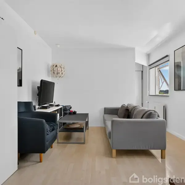 Stue med grå sofa og sort lænestol står ved et glasbord. Et TV er placeret på skænken. Væggene er hvide, og store vinduer lader lys ind.