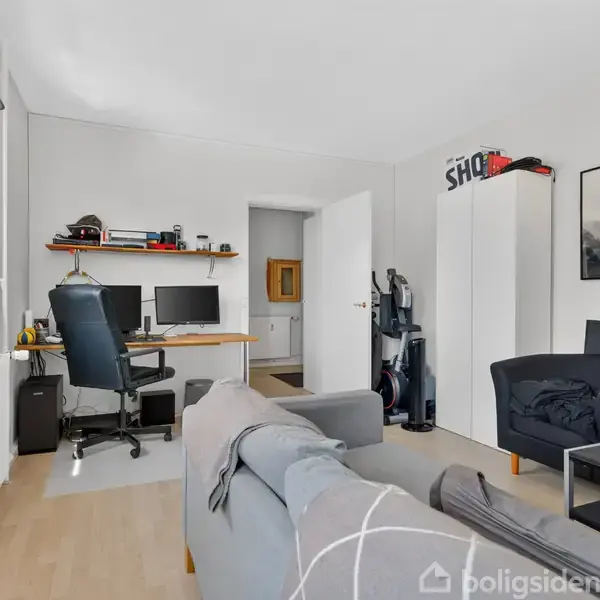 Stue med en sofa og skrivebord, hvor to skærme står. En kontorstol foran skærmene. Reoler og skabe omkranser rummet, der er lyst op af et vindue.