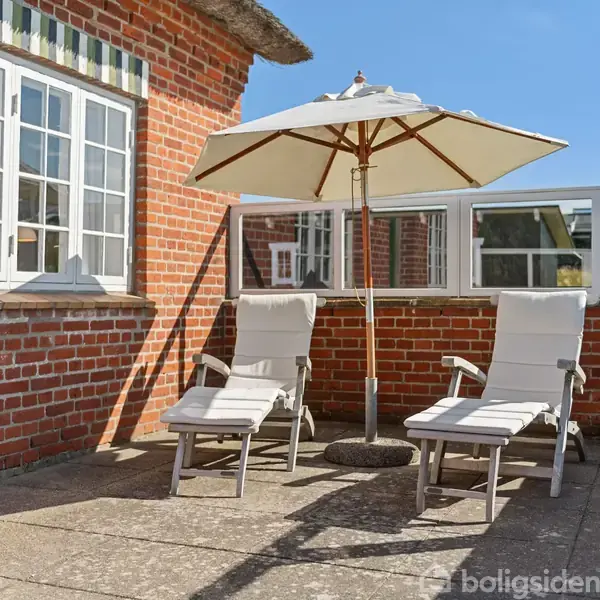 To hvide liggestole står under en parasol på en solbeskinnet terrasse omgivet af røde murstensvægge med store vinduer.