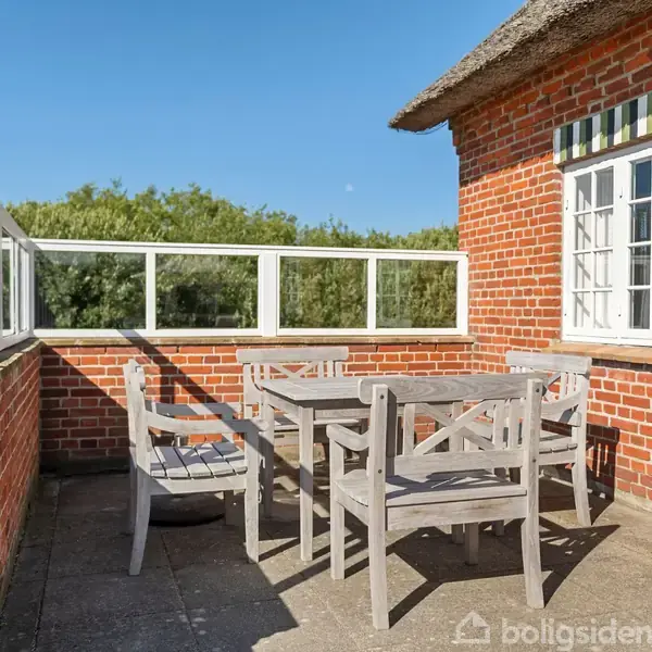 Træmøbler står på en terrasse, omgivet af murstensvæg og glasrækværk. Ved siden af bygningen med hvidrammet vindue og stribet markise, skaber det en hyggelig udendørs plads under blå himmel.