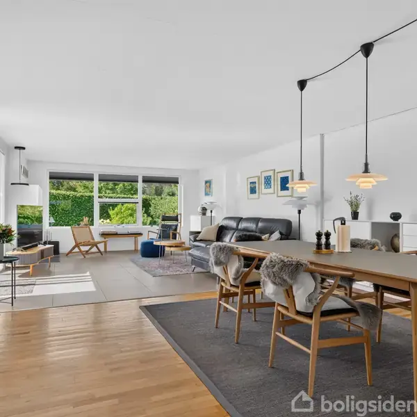 Stort, lyst stueområde med spisebord og stole i forgrunden, sofa og sofaborde i baggrunden. Vinduer åbner til grøn udsigt. Trægulv og moderne lamper i loftet.