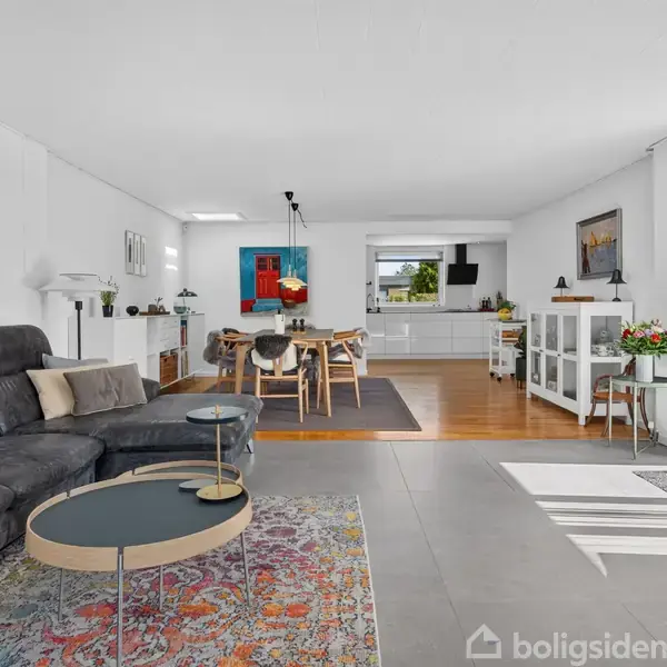 Sort lædersofa står i en lys stue med farverigt tæppe og runde sofaborde foran. Spisebord og stole ses bagerst, med køkken i baggrunden. Store vinduer til højre.