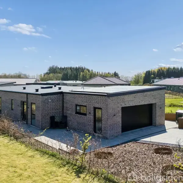 En moderne, lav murstensbungalow står stille i et åbent boligområde med græsplæne og omkransende buske under en blå himmel med spredte skyer.