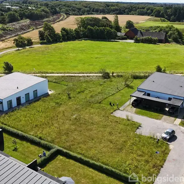 To moderne huse står side om side, adskilt af et åbent grønt græsareal. Omkring dem er der marker og træer, hvilket skaber en landlig atmosfære.