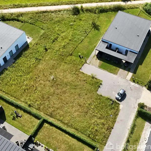 Et ubebygget grundareal med vildtvoksende græs ligger mellem to moderne, firkantede huse med flade tage. En bil holder parkeret foran et af husene på en asfaltvej.