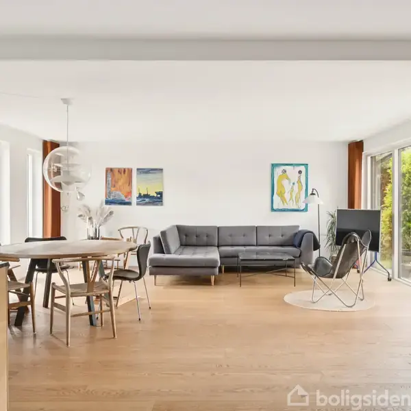 Stue med grå sofa i hjørnet, tv på stativ, og spisebord ved siden af. Store vinduer giver udsigt til en grøn have. Enkel indretning med kunst på væggene.