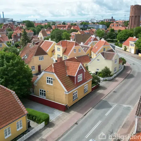 En række gule huse med røde tegltage langs en vej. Træer omgiver området, og i baggrunden ses industribygninger og en høj vandtank.