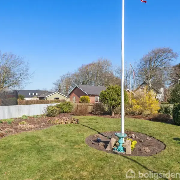 Flagstang står i en velholdt have med grøn græsplæne og buske. I baggrunden ses trækroner og hustage under en klar blå himmel.