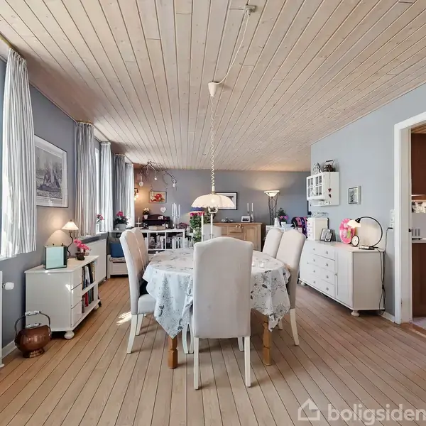 Et spisebord med hvide stole er centralt placeret i en lys stue. Rummet har trægulv, hvide vægge og forskellige møbler, herunder kommoder og reoler. Vinduer lader dagslyset strømme ind.