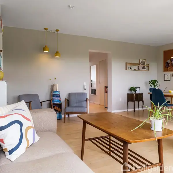 Beige sofa med farverig pude, står ved træbord. Rum indeholder grønne planter, stole, hylde med dekoration og spisebord i lyse stue.
