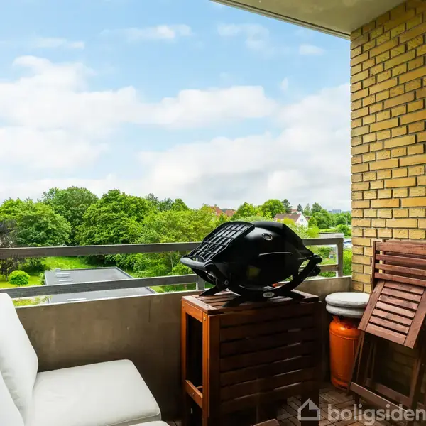 En balkon indeholder en hvid sofa, en grill placeret på et lille bord, og sammenfoldelige stole ved siden af. Omgivet af gule murstensvægge og udsigt til en grøn have.