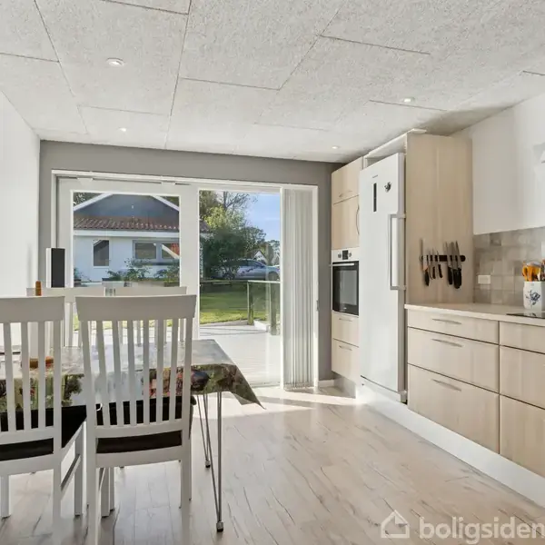 Køkken-spisestue med lang bordplade, træskabe, og spisebord med to stole. Stort vindue i baggrunden giver udsigt til terrasse og have.