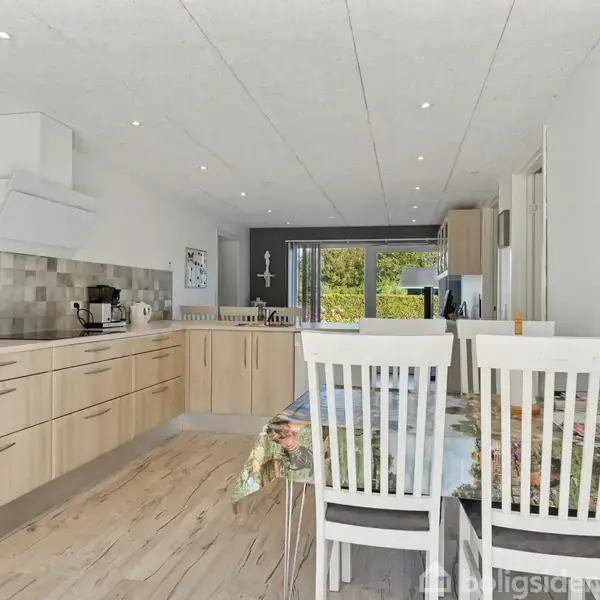 Et lyst køkken med træskabe og bordplader, hvor en kaffemaskine og frugtskål står. Spisebord med blomstret dug og hvide stole. Store vinduer giver udsigt til grøn have.