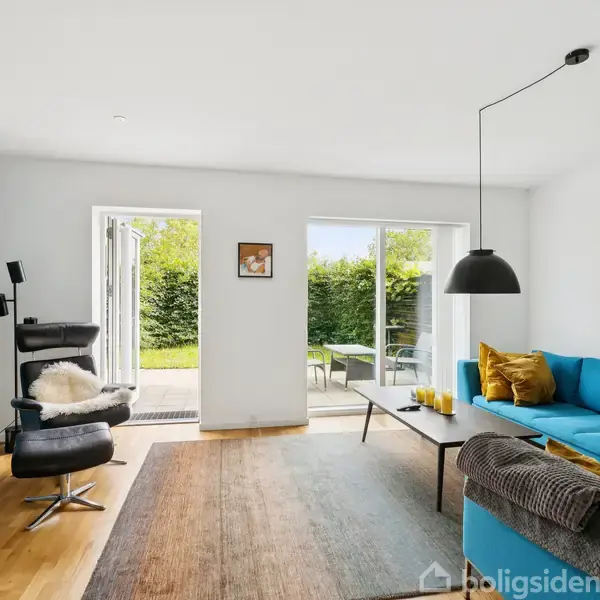 En blå sofa står i stuen, omgivet af moderne møbler som en lænestol og et sofabord. Gennem de åbne terrassedøre ses en have med hække. Over sofaen hænger en sort-hvid væglampe.