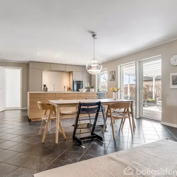Et lyst, moderne køkken-alrum med et spisebord og stole i centrum. Rummet har en glashylde til venstre, store vinduer mod en terrasse, og en åben dør til et tilstødende rum. En pendellampe hænger over bordet.