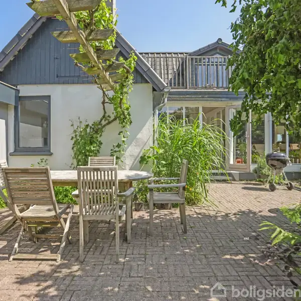 En trægårdsplads med træmøbler står på en brolagt terrasse ved siden af grøn beplantning. I baggrunden ses et hus med store vinduer og en balkon, belyst af solskin.