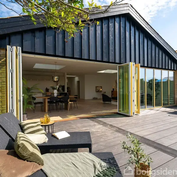 Et moderne hus med åbne glasfoldedøre fører fra en stue til en stor træterrasse. Terrassen er udstyret med udendørs møbler og omgivet af hegn og planter.