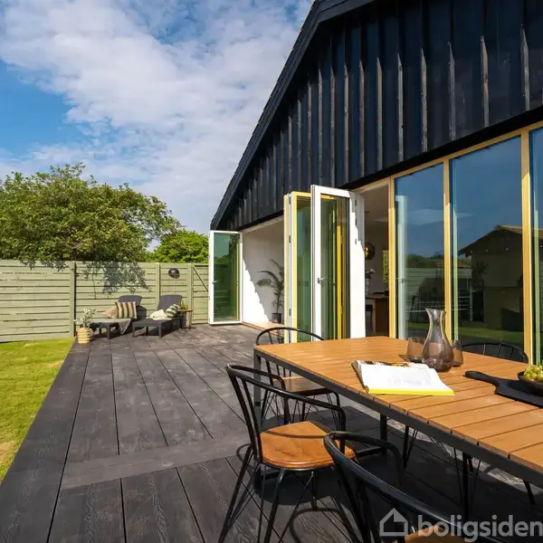 En træterrasse med bord og stole i forgrunden, åbne døre til et moderne hus i mørkt træ, omkranset af en grøn have og stakit under en blå himmel.