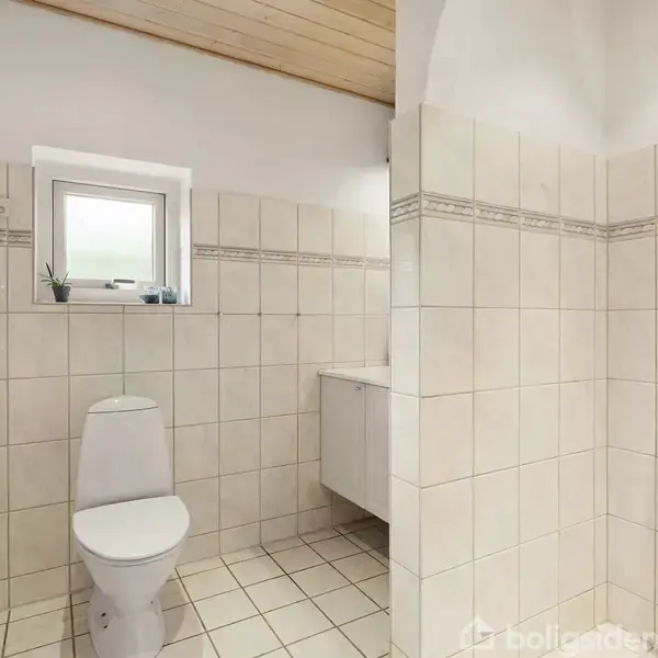 Toilet står stille i hvidt flisebelagt badeværelse med bruser til højre. Lille vindue over toilettet, hvidt loft, og håndklædetørrer til venstre.