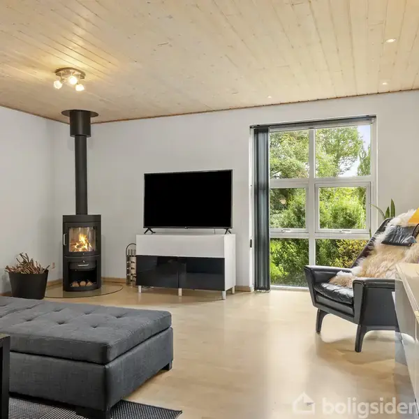 Stue med sort brændeovn i hjørnet, fladskærms-TV på hvid stand, glasdør med udsigt til have og sofa samt lænestol med gulvlampe. Lys indretning.