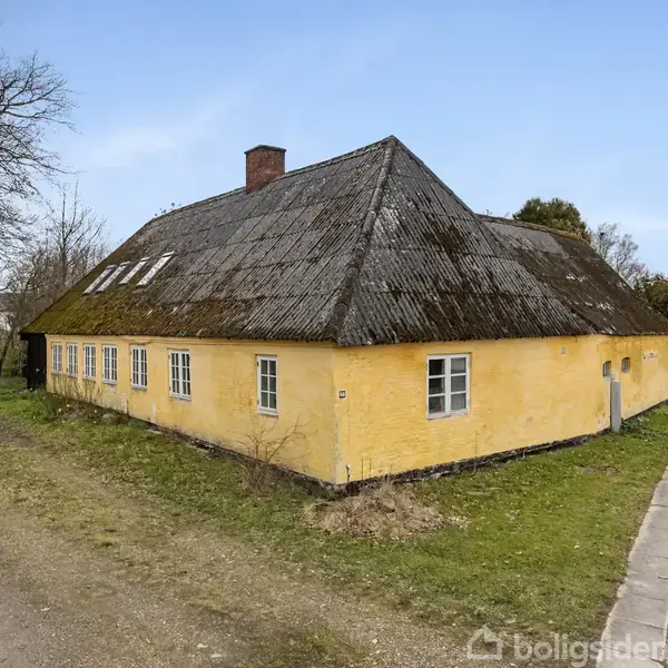 Gult, traditionelt hus med mørkt, skråt tag står ved en vej. Der er flere små vinduer langs væggene. Omgivet af græs og træer.