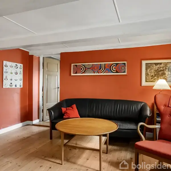 En stue med røde vægge indeholder en sort lædersofa, en rund træsofabord og en rød lænestol. Lamper og kunst hænger på væggen, hvilket skaber en hyggelig atmosfære.