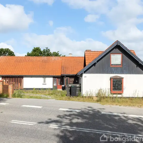 Et hvidt hus med rødt tag står ved vejen. En grå postkasse og en hæk ses foran. Bagved er der træer og en blå himmel med skyer.