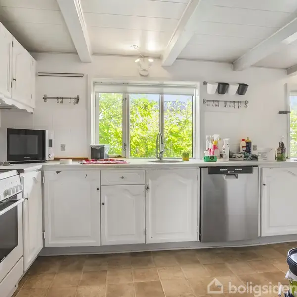 Køkken med hvide skabe og træloft, hvor opvaskemaskinen og en mikroovn er placeret. Vinduet åbner mod grønne træer, og der findes køkkenudstyr og rengøringsmidler på bordpladen.