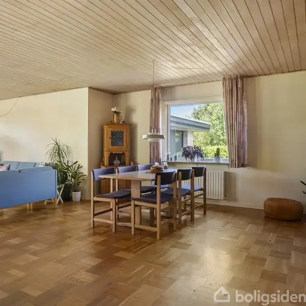 En spisestue med træbord og seks stole. Ved siden af ses stue med blå sofa. Store vinduer og planter skaber en lys atmosfære. Parketgulv og lyst træloft.