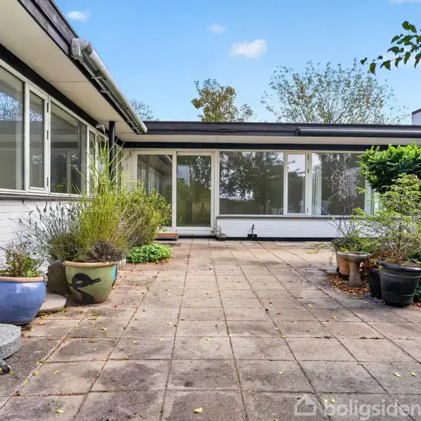 Stenterrasse med blomsterkrukker i forskellige størrelser, placeret foran et moderne, hvidt hus med store vinduespartier. Terrasse er omgivet af frodig beplantning og enkelte træer.