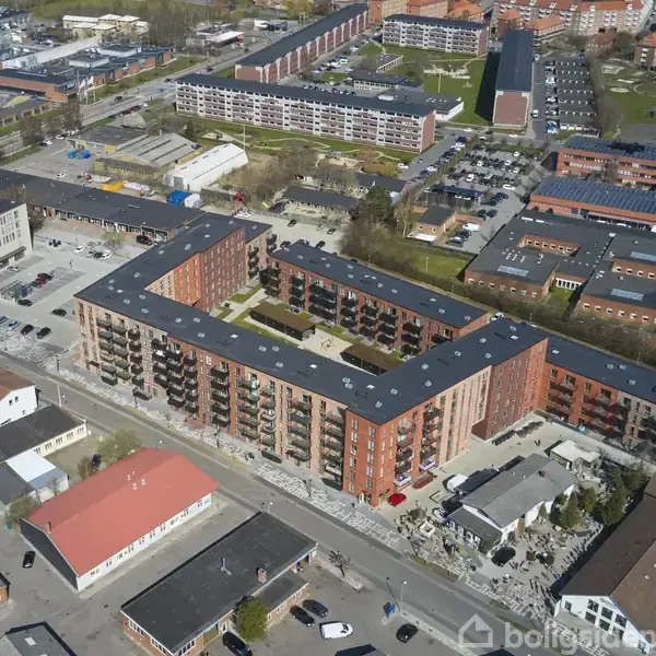 Et stort, L-formet boligkompleks i røde mursten dominerer billedet, omgivet af mindre bygninger og parkeringspladser, beliggende i et byområde med flere lignende komplekser i baggrunden.