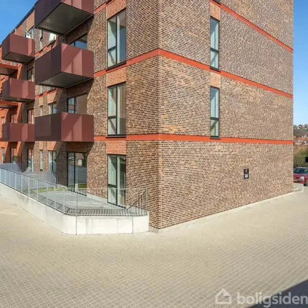 Bygning med røde murstensfacader og tilhørende altaner strækker sig langs en flisebelagt gade. Parkeringsplads og få træer ses i baggrunden under en blå himmel.