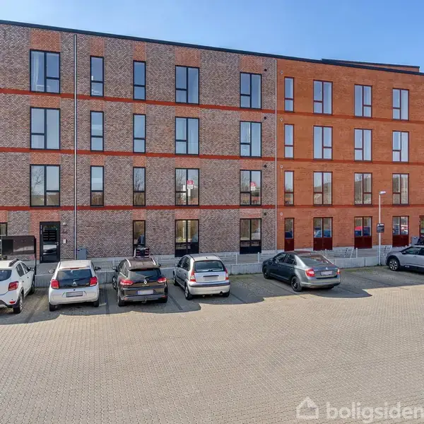 Bygning med tre etager har flere vinduer og rødbrune mursten. Foran er biler parkeret på en åben parkeringsplads med fliser.