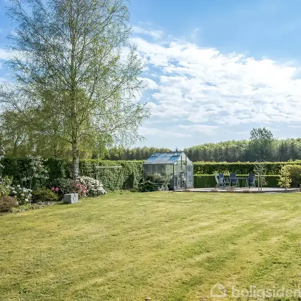 Drivhus står stille ved siden af et bord med stole på en terrasse i en velplejet have, omgivet af grønne buske og træer under en blå himmel med spredte skyer.