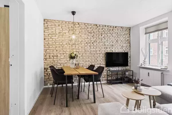 Et spisebord med fire stole står foran en murstensvæg i en stue. En lampe hænger over bordet, og en fladskærm er placeret på en reol ved siden af et vindue.