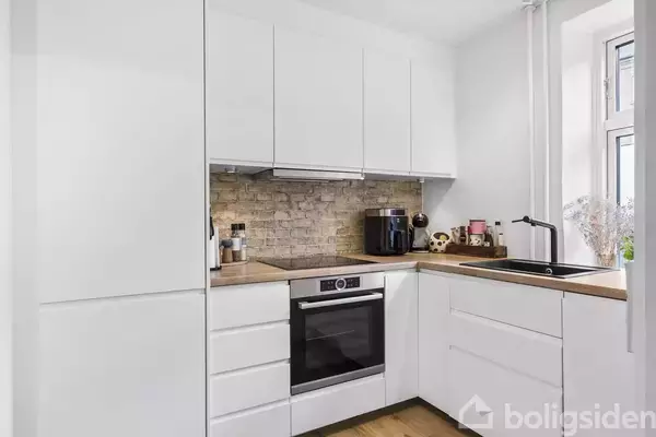 Hvidt køkken med moderne elementer og træbordplade. En ovn og kaffemaskine står på bordet ved en murstensvæg. Et vindue giver lysindfald ved vasken.