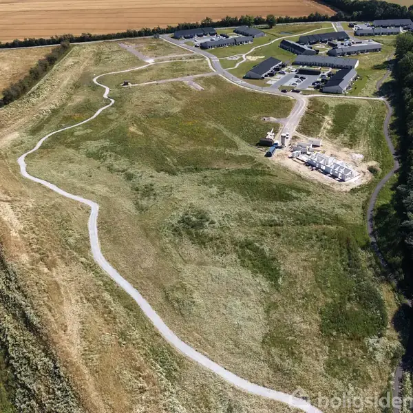 Stort grønt område med snoet sti, omgivet af marker. Huse og byggeri ses i udkanten af området. En stille landskabsscene, der indgyder ro.