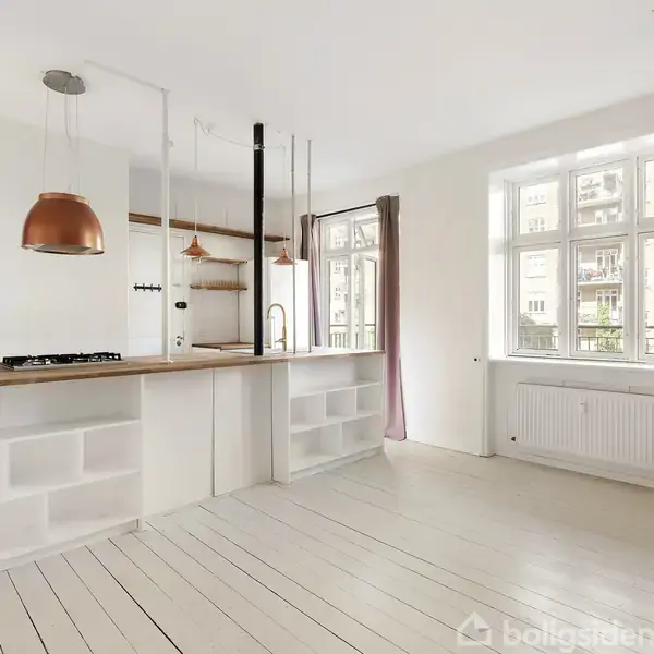 Et lyst køkken med åben planløsning, højbord med trækogeplade og kobberlamper. Store vinduer lader naturligt lys strømme ind, mens hvide vægge og gulve skaber en rummelig atmosfære.