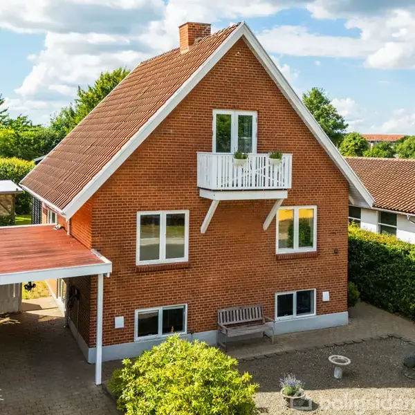 Rødt murstenshus med hvidt tag og balkon, omgivet af en have og hæk. Bilenport ved siden af huset. Blå himmel med skyer i baggrunden.