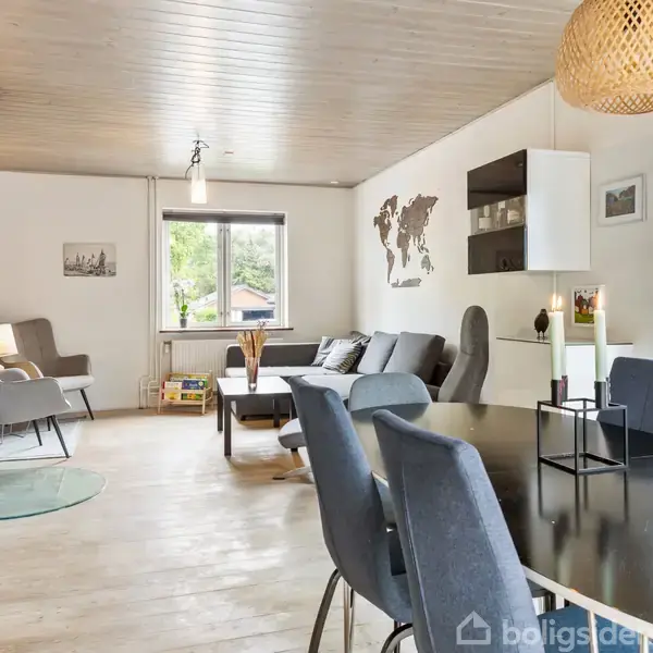 En moderne stue med spisebord og stole i forgrunden. Sofa og lænestole står ved vinduerne i baggrunden. Hængelampe og verdenskort på væggen fuldender indretningen.