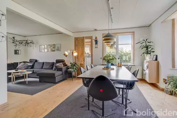 Stue med sort sofa og sofabord til venstre, spisebord med sorte stole til højre. Rummet er lyst med træpaneler, planter og billeder på væggen. Vindue med udsigt udenfor.