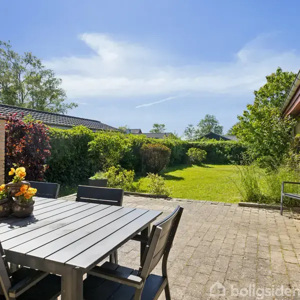 Et sort spisebord med stole er placeret på en flisebelagt terrasse ved siden af et hus i gule mursten. I baggrunden ses en grøn have med buske og træer.